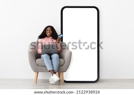 Mobile shopping application. Happy black lady sitting on chair near big cellphone with empty screen using credit card and laptop, white studio wall background, mockup, collage Royalty-Free Stock Photo #2122938926