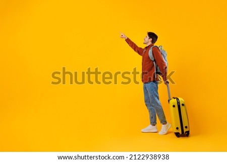 Happy Tourist Man Pointing Finger Aside Standing With Travel Suitcase And Backpack Posing Over Yellow Studio Background. Tourism And Vacation Offer Concept. Full Length, Empty Space For Text Royalty-Free Stock Photo #2122938938