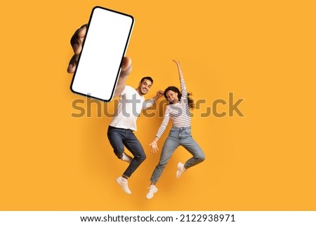 Wow, great news. Overjoyed couple showing huge smartphone with blank white screen, jumping and celebrating victory over orange studio background, mockup collage