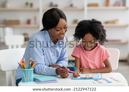 Cute little black girl with bushy hair exercising at daycare, sitting at table with friendly female teacher and doing various tests, young woman psychologist working with child preschooler Royalty-Free Stock Photo #2122947122