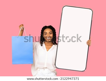 Shopping application concept. Happy african american lady showing big smartphone with empty screen and holding shopper bag standing on pink studio background, mockup