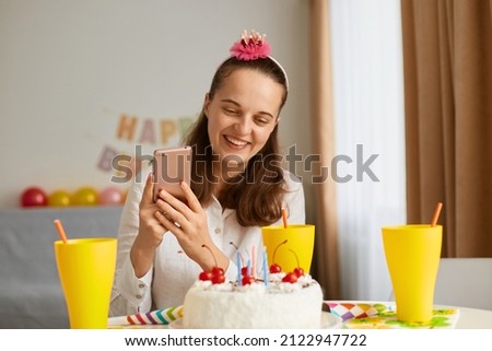 Portrait of smiling happy young woman wearing white shirt celebrating birthday alone, holding mobile phone in hands, taking photo of her holiday dessert, expressing positive emotions.