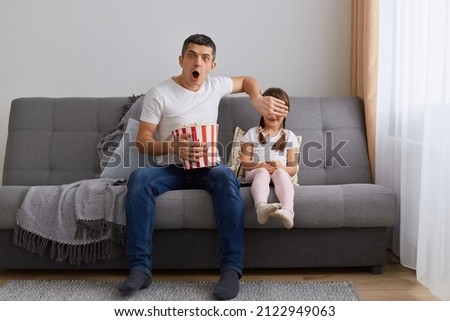 Portrait of attractive shocked father watching horror film at home with little daughter, covering her eyes with his palm, eating popcorn, man keeps mouth open.