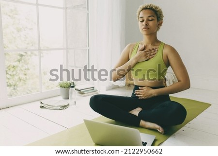 African female yoga teacher having online lesson sitting on green mat in light room showing pranayama techniques, hands on her chest and belly, looking concentrated and focused on body feelings Royalty-Free Stock Photo #2122950662