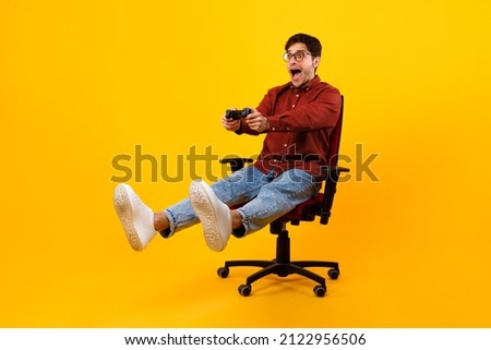 Emotional Male Gamer Shouting Playing Videogame Sitting In Chair And Holding Gamepad Controller Looking Aside Over Yellow Studio Background. Computer Gaming Concept