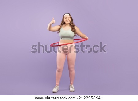 Satisfied young caucasian plus size lady athlete in sportswear with hula hoop show thumb up isolated on purple background, studio. Approve training, body care and weight loss. Fit blog and workout