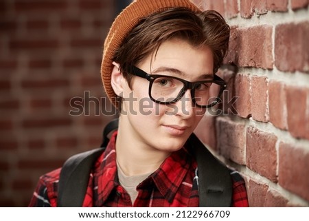 Youth lifestyle. A handsome joyful modern teenage boy in glasses and stylish casual clothes standing leaning against a brick wall. Carefree adolescence. 
