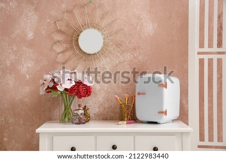 Small cosmetic refrigerator, makeup brushes and vase with flowers on table near color wall