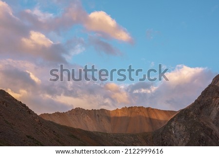 Dramatic landscape with sunlit wide sharp mountain ridge under clouds in sunset colors at changeable weather. Atmospheric mountain scenery with large sharp rocks on ridge top under sunset cloudy sky.
