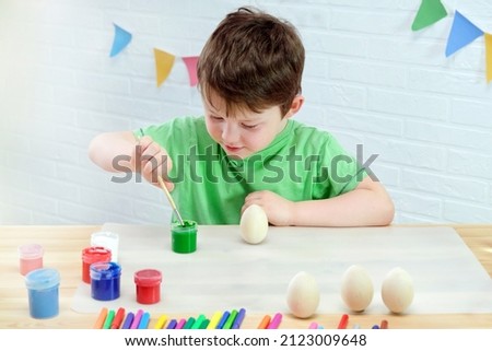 a boy of 4-5 years old sits at a table in a room and paints wooden Easter eggs with green paint. A child in a green T-shirt against a white brick wall. Creative occupation, preparation for Easter. Royalty-Free Stock Photo #2123009648