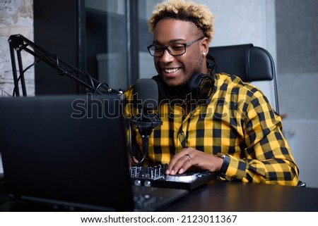 Positive African American DJ is working on digital mixing in a home recording studio. A black man with a fashionable hairstyles uses a small DJ console, a microphone and a laptop to record a new track