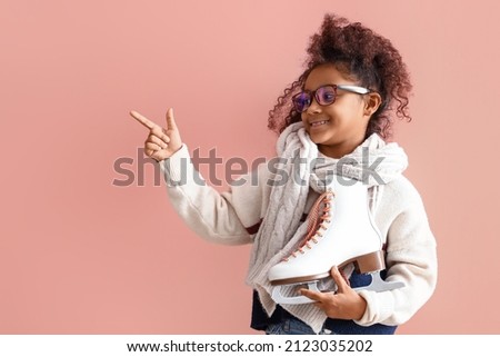 Little African-American girl in eyeglasses with ice skates pointing at something on pink background