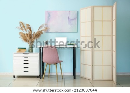 Modern workplace with laptop, chest of drawers and folding screen near blue wall