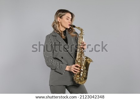 Beautiful young woman in elegant suit playing saxophone on grey background