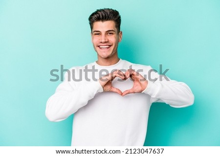 Young caucasian man isolated on blue background smiling and showing a heart shape with hands.