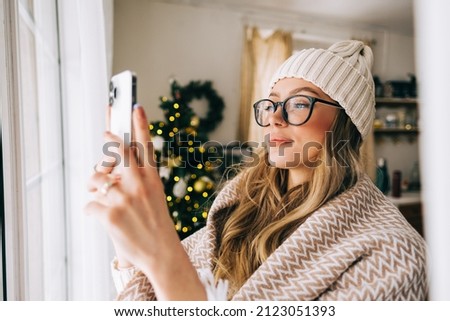 Young caucasian woman standing near window at home and taking photo after snowfall in the morning.