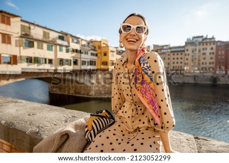Young woman enjoys beautiful view on famous Old bridge in Florence, sitting on the riverside at sunset. Female traveler visiting italian landmarks. Stylish woman wearing dress and colorful shawl Royalty-Free Stock Photo #2123052299