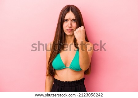 Young caucasian woman wearing bikini isolated on pink background showing fist to camera, aggressive facial expression.