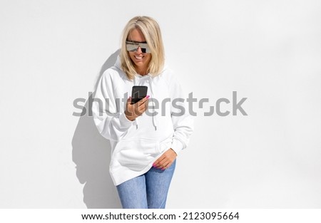 Woman in white hoodie standing by white wall and using mobile phone