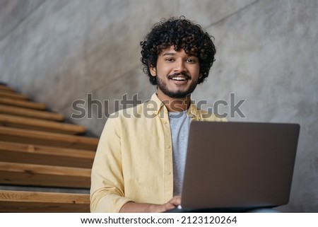 Handsome Indian businessman using laptop computer working in modern  office. Asian student studying, learning language, online education concept. Smiling freelancer copywriter typing looking at camera