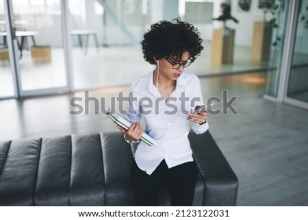 Millennial caucasian businesswoman listening music in earphones and using smartphone. Concept of modern successful woman. Young focused curly girl sitting on leather bench at open space office