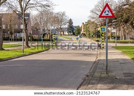 Triangular traffic sign for roundabout, pedestrian crossing, metal post with a triangular plate in red, black, white and arrows, roundabout and house in the blurred background, Netherlands