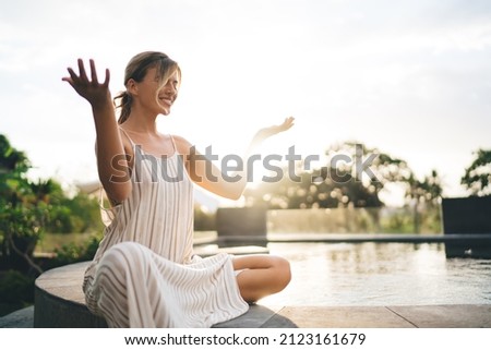 Young happy woman practicing yoga and meditating on swimming pool edge in yard of resort hotel. Tourism, vacation and weekend. Pretty caucasian girl. Sunset. Idyllic and calm lifestyle on Bali island