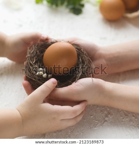 Happy Easter. Kids' hands hold Easter eggs in a small nest on a white table. Egg decoration, festive cooking. Christian tradition. Square photo