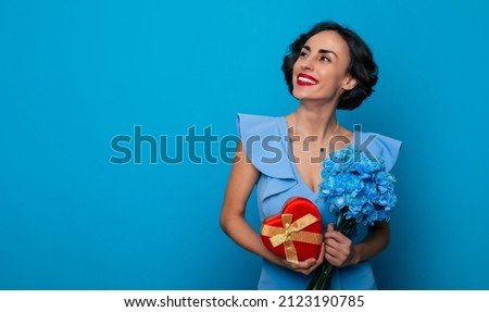 Women's day. St. Valentine's day. Close up portrait of elegant fashionable smiling woman in dress with bunch of flowers and gift box in hands isolated on blue background