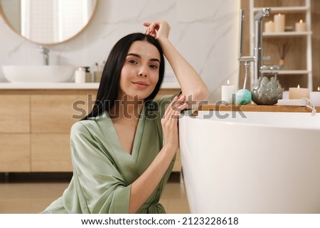 Beautiful young woman sitting near tub in bathroom