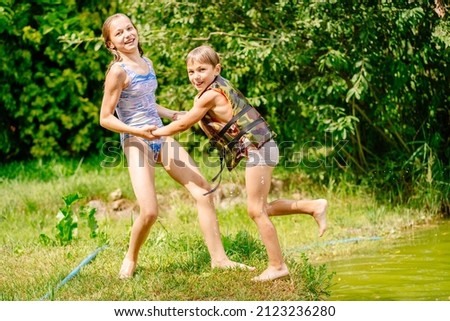 Cute blond preteen sister helping her little brother out of the water. Children in swimsuit plays together. Summer, holiday, outdoor activities at summer time concept.