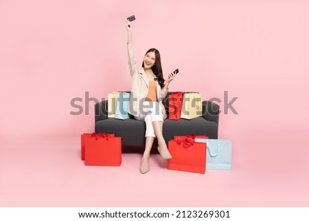 Full length portrait of Excited Asian woman sitting on sofa with shopping bags and hands up with credit card and mobile phone isolated on pink background, Shopper or shopaholic concept Royalty-Free Stock Photo #2123269301