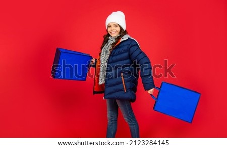 happy kid in puffer jacket and hat. teen girl after shopping on red background.