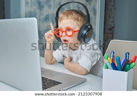Cute child in red glasses and headphones has fun communicating online via video link using a laptop. The concept of modern means of communication and communication at a distance