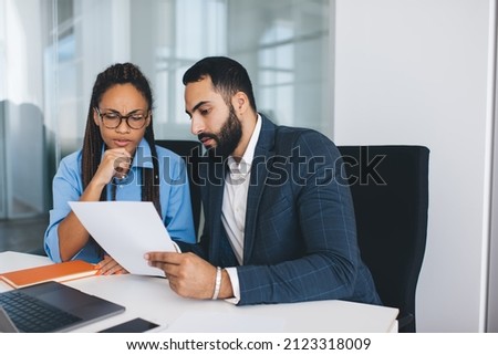 Puzzled male and female colleagues discussing information during cooperation briefing in office interior, multicultural partners in formal clothes analyzing paperwork reports and documents in company Royalty-Free Stock Photo #2123318009