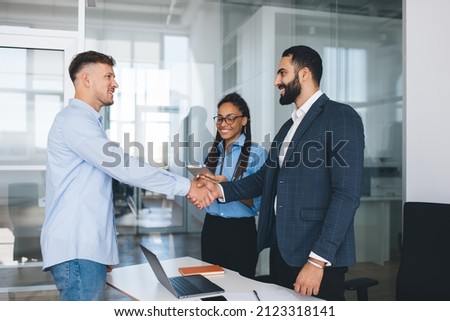 Multiracial male employees in smart casual clothing doing handshake during business meeting in office interior during recruitment interview, proud ceo found merger agreement of acquisition on deal Royalty-Free Stock Photo #2123318141