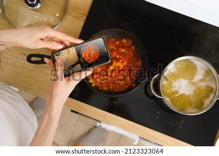 View from above of unrecognizable woman with smartphone taking photo of frying pan with tomato sauce for culinary blog while cooking in kitchen at home. Food blogger recording video of cooking process