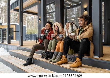 Group of young friends sitting on stairs outside the country house, drinking coffee and enjoying the winter