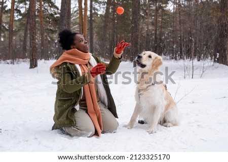 African woman in warm clothing playing ball with her dog during their walking in the forest in winter