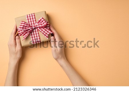 First person top view photo of saint valentine's day decorations female hands taking craft paper giftbox with checkered ribbon bow on isolated beige background with empty space