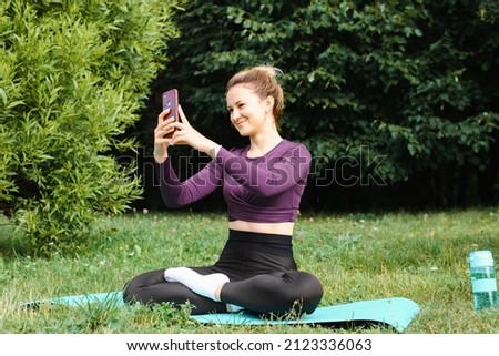 Pretty fit young woman with cute smile taking sefli on phone while practicing yoga and meditating on lawn in park. Slim sportswoman training on sports mat and posting pictures in social media outdoors