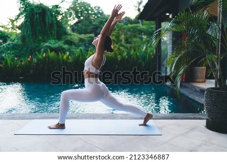 Sportive woman dressed in white tracksuit doing yoga practice for feeling bliss at patio terrace with pool, slim female checking body flexibility while exercising during holistic retreat healing