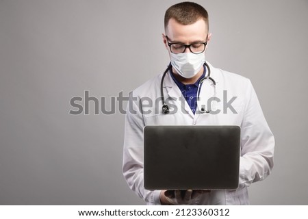 young caucasian doctor in a mask and glasses uses his laptop. studio portrait