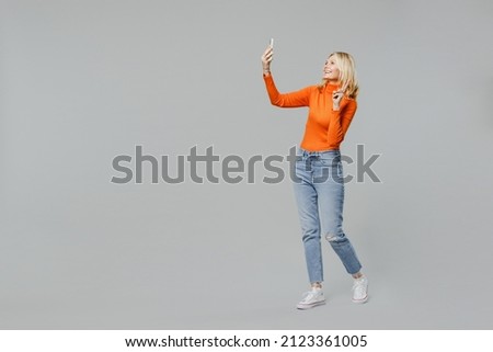 Full body elderly smiling happy blonde woman 50s in orange turtleneck doing selfie shot on mobile cell phone post photo on social network show v-sign isolated on plain grey background studio portrait.