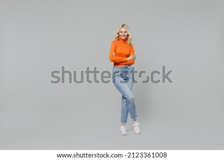 Full body elderly smiling happy blonde caucasian woman 50s in orange turtleneck hold hands crossed folded look camera isolated on plain grey color background studio portrait. People lifestyle concept