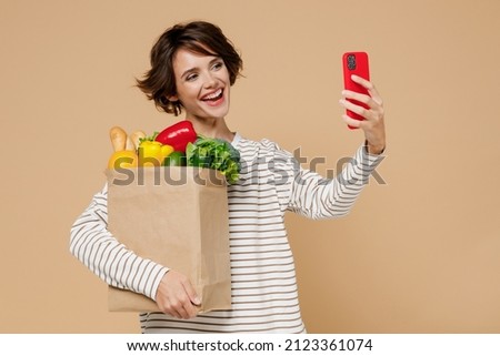 Young smiling woman 20s in casual clothes hold paper bag with vegetables doing selfie shot on mobile cell phone post photo on social network isolated on plain pastel beige background Shopping concept