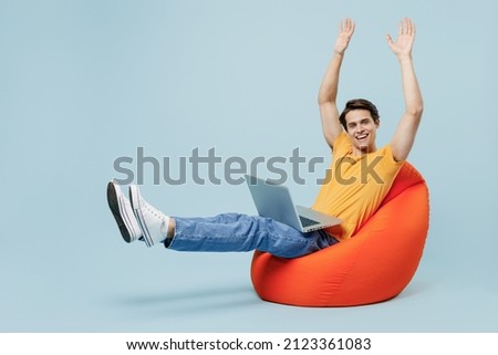 Full body side view fun young man 20s wearing yellow t-shirt sit in bag chair hold use work on laptop pc computer stretch hands legs finish legs isolated on plain pastel light blue background studio.