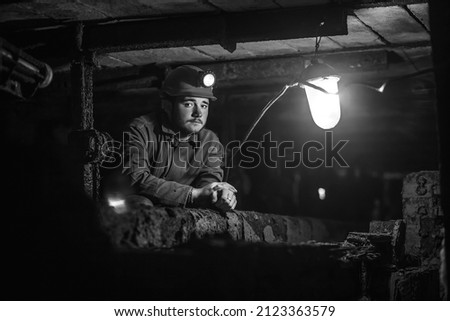Tired young miner in an old coal mine. A miner in a protective suit with a helmet on his head and a flashlight works in the dark. Work in a coal mine.  Royalty-Free Stock Photo #2123363579