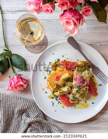 Gourmet white fish recipe with assorted citrus fruits, olives, fresh herbs, marinated red onion and fennel, served on a white ceramic plate and with a glass of white wine. Table decoration with roses Royalty-Free Stock Photo #2123369621