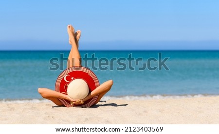 A slender tanned girl on the beach in a straw hat in the colors of the flag of Turkey. The concept of a perfect vacation in a resort in the Turkey. Focus on the hat. Royalty-Free Stock Photo #2123403569